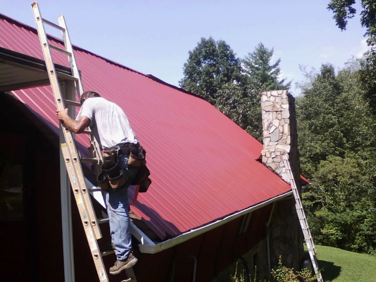 Roofer on a ladder beside a red metal roof and stone chimney — Soffit & Fascia Repair by Roofing Brothers in Weirsdale